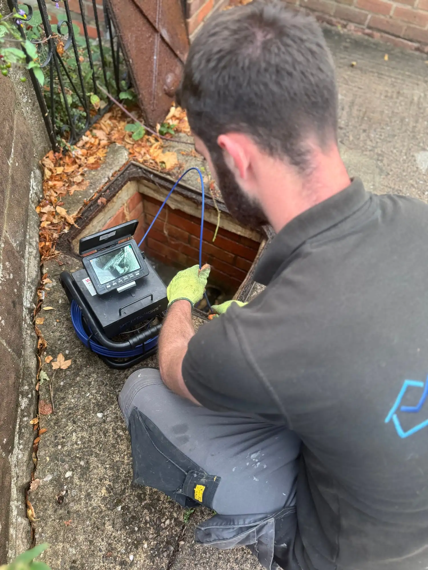 Noah kneeling at an open inspection chamber, feeding a drain survey camera in, with the camera screen showing the inside of the pipe